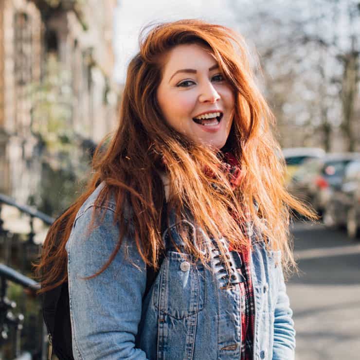 Ashley Baxter wearing a jeans jacket and backpack and laughing into the camera